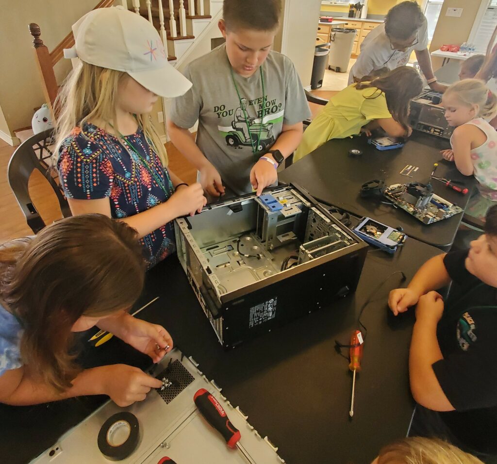 Students gather around a deconstructed computer, surrounded by parts and tools. 