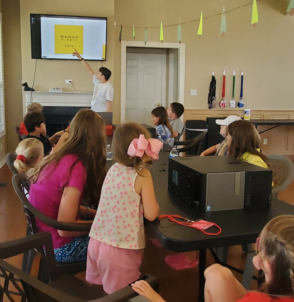 Students sit around a table with a desktop computing lying on it, listening to a presentation. 