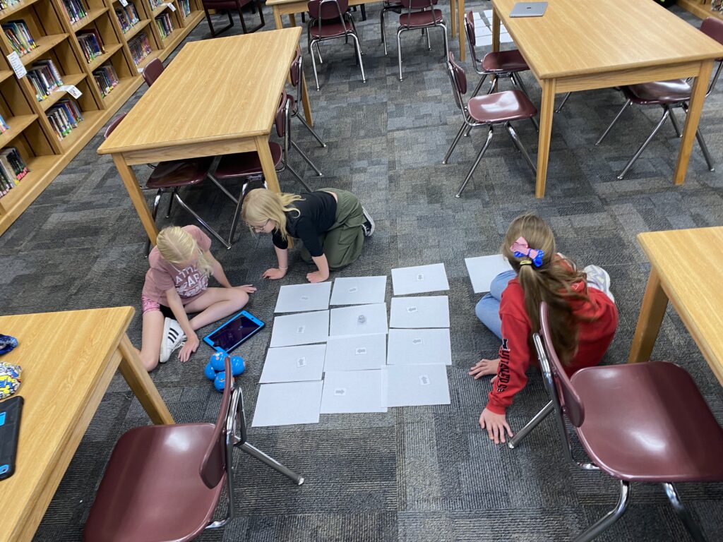 Three students sit on a carpeted floor with a Dash robot, tablet, and various papers in a square. 