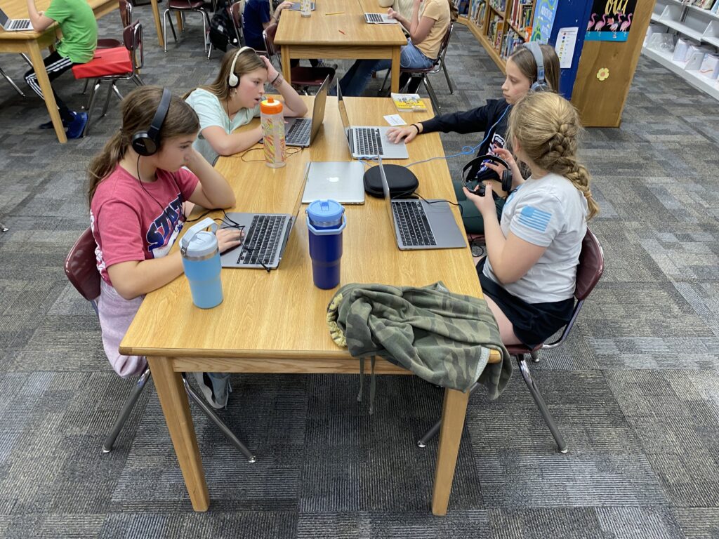 Students sit on laptops and headphones around a table. 