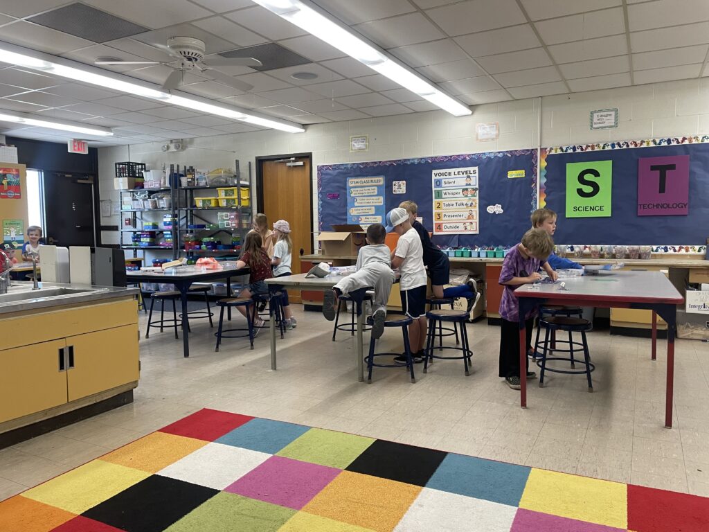 Students gather around tables in a classroom, exploring with and assembling Snap Circuits. 