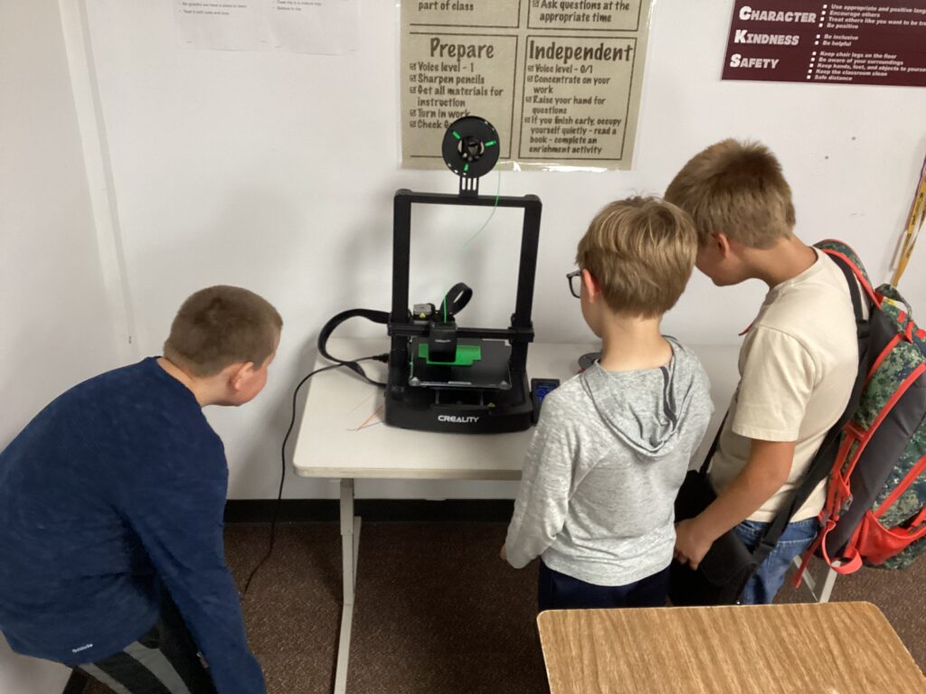 Three students gather around their 3D printer, watching a print in action. 