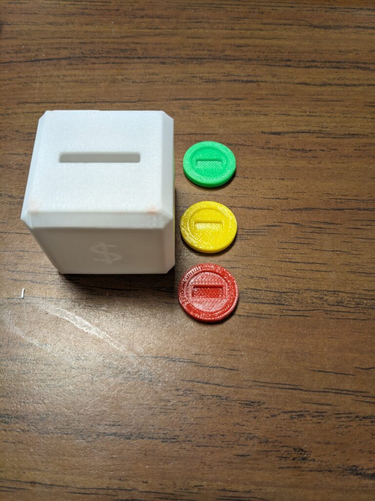 The 3D printed product of a small box with a slot on top and a money sign, with three coins next to it -- green, yellow, and red. 