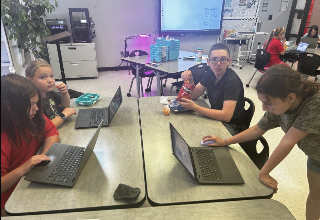 Students sit around a classroom table, each working on a laptop. 