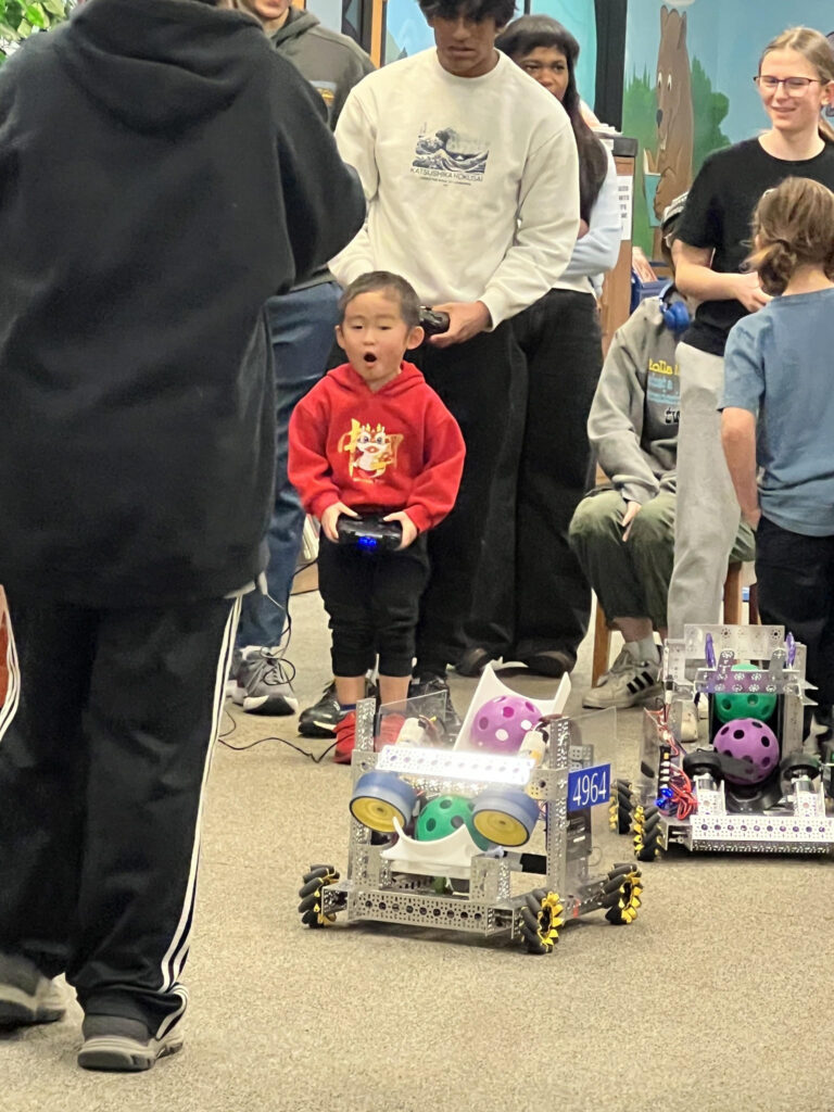 A photo from Rolla Public Library's robotics event, showing a group of people standing around a robot with one child, a look of amazement on his face, controlling it. 