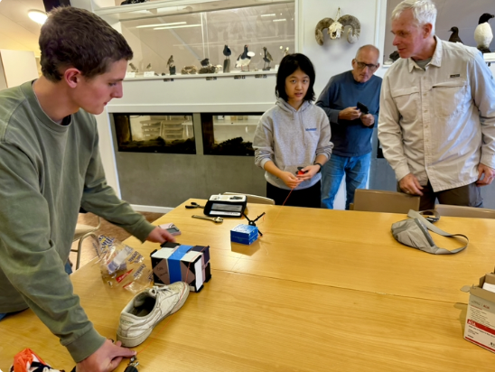 Individuals gather around a table, working on assembling their experiment prior to the balloon launch. Pictured on the table are various boxes, tape pieces, and a sneaker.