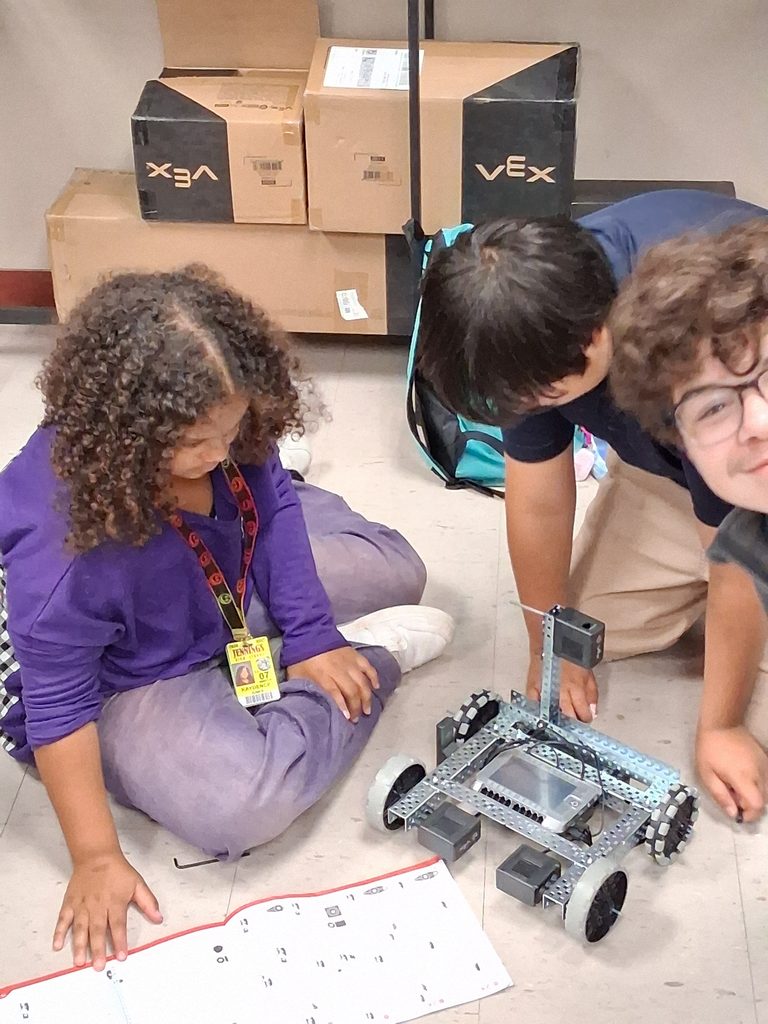 Robotics team members gather on a classroom floor, surrounding VEX equipment.