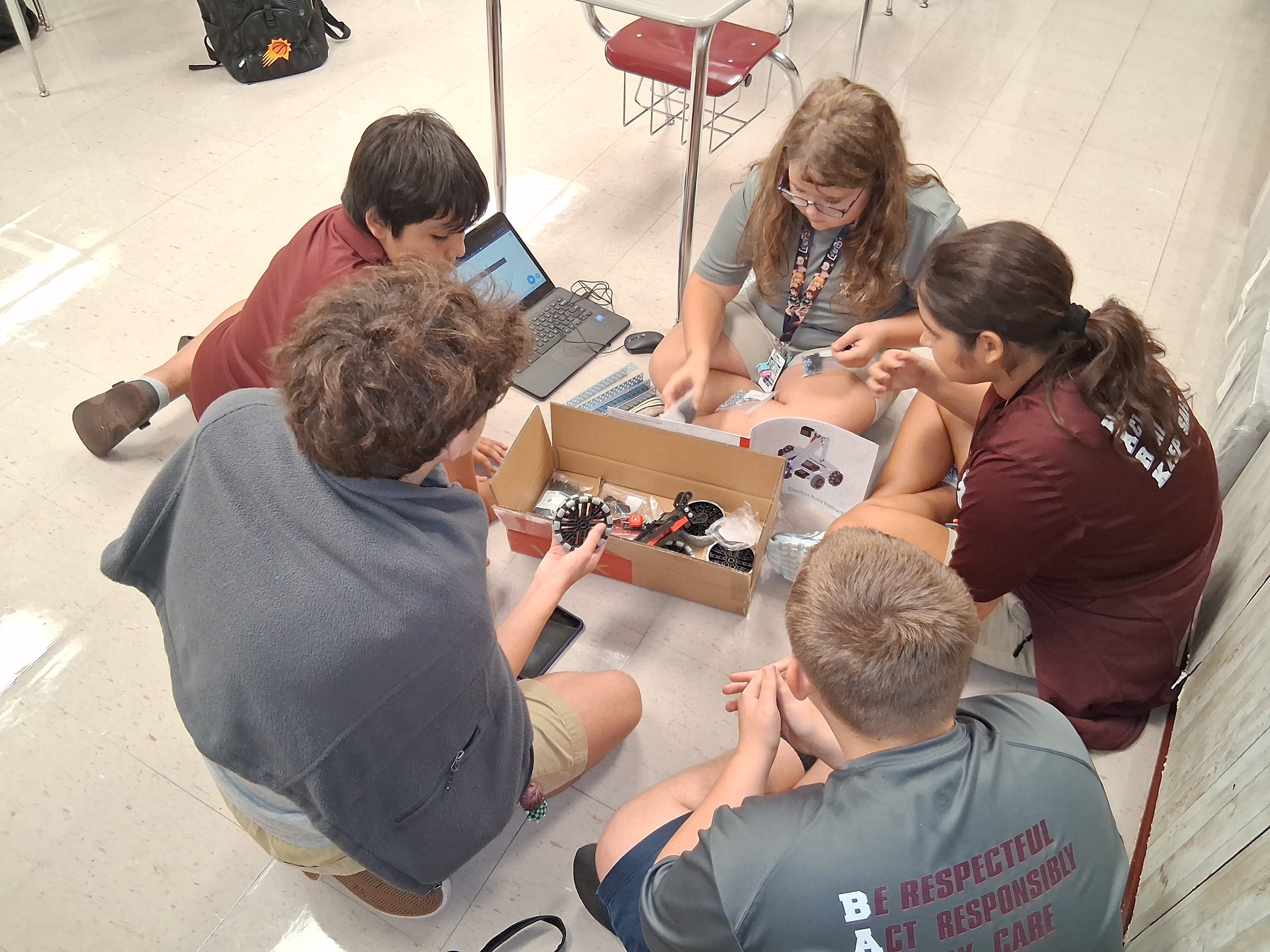 The robotics team gathers in a circle on a classroom floor, surrounding VEX equipment and a laptop.