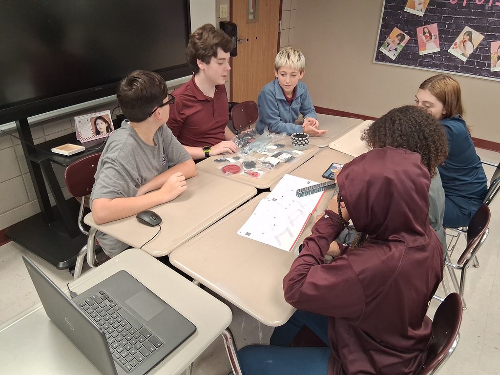 Robotics team members gather in a table made of desks, surrounding various packages of VEX equipment and a manual.